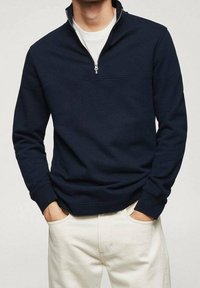 Sudadera con cremallera en color navy, fabricada con un tejido suave, con un cuello levantado, cremallera corta y puños acanalados. Combinada con pantalones de color claro.