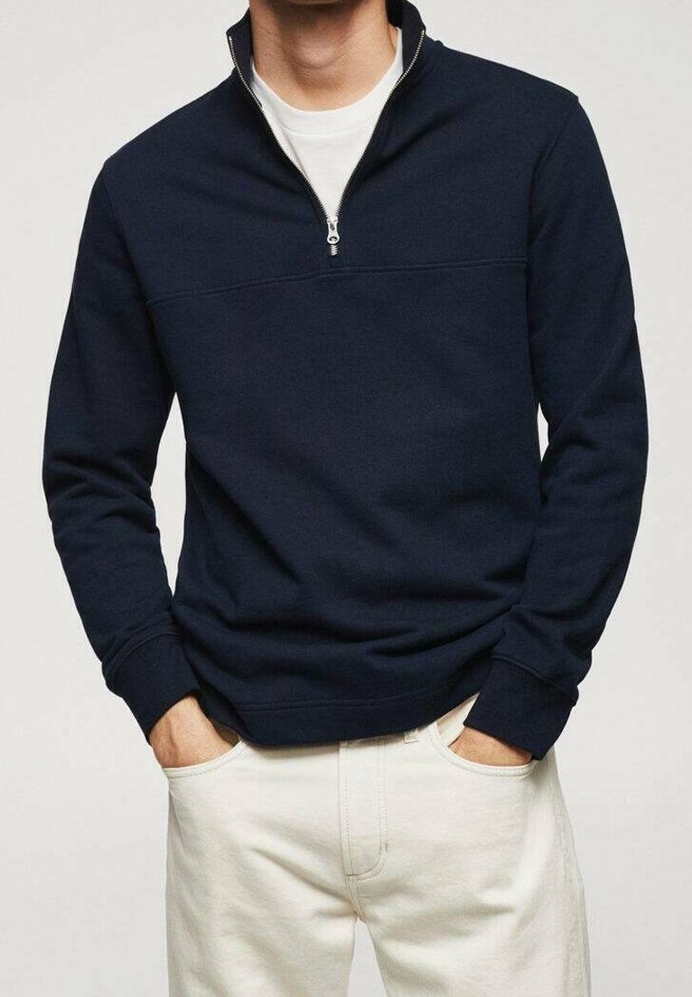 Sudadera con cremallera en color navy, fabricada con un tejido suave, con un cuello levantado, cremallera corta y puños acanalados. Combinada con pantalones de color claro.