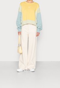 Persoon in een geel, roomkleurig en blauw kleurblok sweatshirt, met roomkleurige wijde broek, witte sneakers, en een kleine gele handtas.