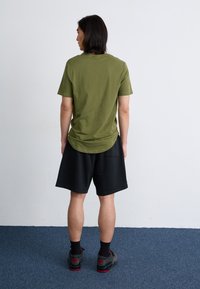 Un jeune homme se tient de dos, portant une chemise verte et un short noir, sur un tapis bleu contre un mur blanc uni.
