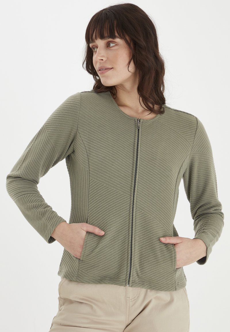 Šviesiai žalia zip-up striukė su grioveliu, ilgomis rankovėmis ir šoninėmis kišenėmis. Medžiagos dizaine yra geometrinės apdailos.