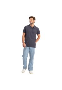 Donkergrijze polo met korte mouwen, met een gele logo op de borst. Draagt een lichte spijkerbroek en witte sneakers.