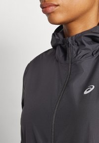 Μαύρο αθλητικό μπουφάν με ψηλό γιακά και φερμουάρ, με ανακλαστικό λογότυπο στο στήθος και λεία, ελαφριά υφή υφάσματος.