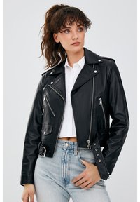 Zwarte leren motorjack met zilveren ritsen, zijzakken en een cropped ontwerp. Gedeeltelijk gedragen over een witte shirt en gecombineerd met hoge taille spijkerbroek.