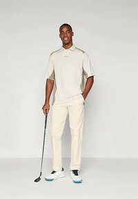 Polo bej de golf cu mâneci scurte și accente deschise, asociat cu pantaloni bej. Persoana ține în mână un ciocan de golf și poartă pantofi albi cu accente albastre.