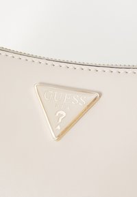 Sac à main beige clair avec un logo Guess triangulaire en or, texte en relief et bords cousus pour un design épuré.