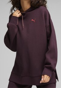 Felpa con cappuccio viola realizzata in tessuto testurizzato. Presenta una tasca frontale a marsupio, cordini con punte metalliche e un piccolo logo rosso sul petto.