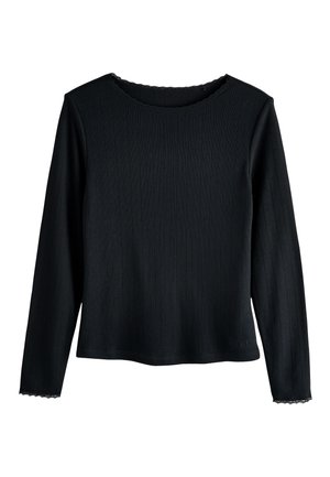 Jeune femme en haut noir à manches longues et pantalon, souriante, debout dans une cuisine moderne avec un plan de travail blanc, des biscuits et une cafetière à piston.