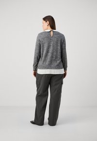 Suéter de punto gris con detalle de abertura en la parte posterior, superpuesto sobre una blusa blanca. Combinado con pantalones cargo gris oscuro. Textura suave.
