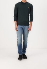 Suéter de punto verde oscuro con cuello redondo y logo amarillo, combinado con jeans de mezclilla azules deslavados y zapatillas negras. Un look simple y casual.
