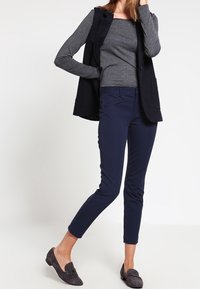 Námořnické slim-fit kalhoty v kombinaci s tmavě šedým dlouhým rukávem a odpovídajícím vestu; ploché černé polobotky dotvářejí outfit. Hladké textury látek.