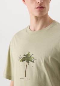 Oliven grøn bomuld t-shirt med et palme-træ grafik i jordnære farver, med teksten "La La La" og "West Coast Stories Since 1993" nedenfor.