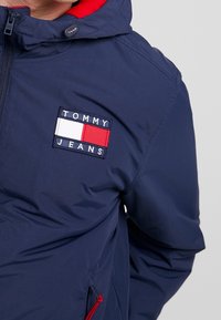 Marineblauer Kapuzenjacke aus leichtem Stoff. Mit rotem Innenfutter, auffälligem gesticktem Logo und verstellbarem Kordelzug am Saum.