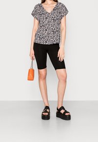 Blouse noire à motif floral avec un col en V, assortie à un short de cycliste noir. Le modèle tient un petit sac orange et porte des sandales plateformes noires épaisses.