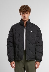 Chaqueta acolchada de puffer negra con cuello alto, logo blanco en el pecho y puños elásticos. Cierre con cremallera en la parte delantera. Material ligero.