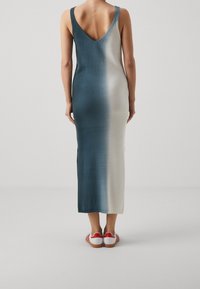 Robe longue sans manches avec un dos en V profond ; dégradé de couleur du vert sarcelle foncé au beige clair ; texture lisse ; fentes latérales ; portée avec des chaussures blanches avec des accents rouges.