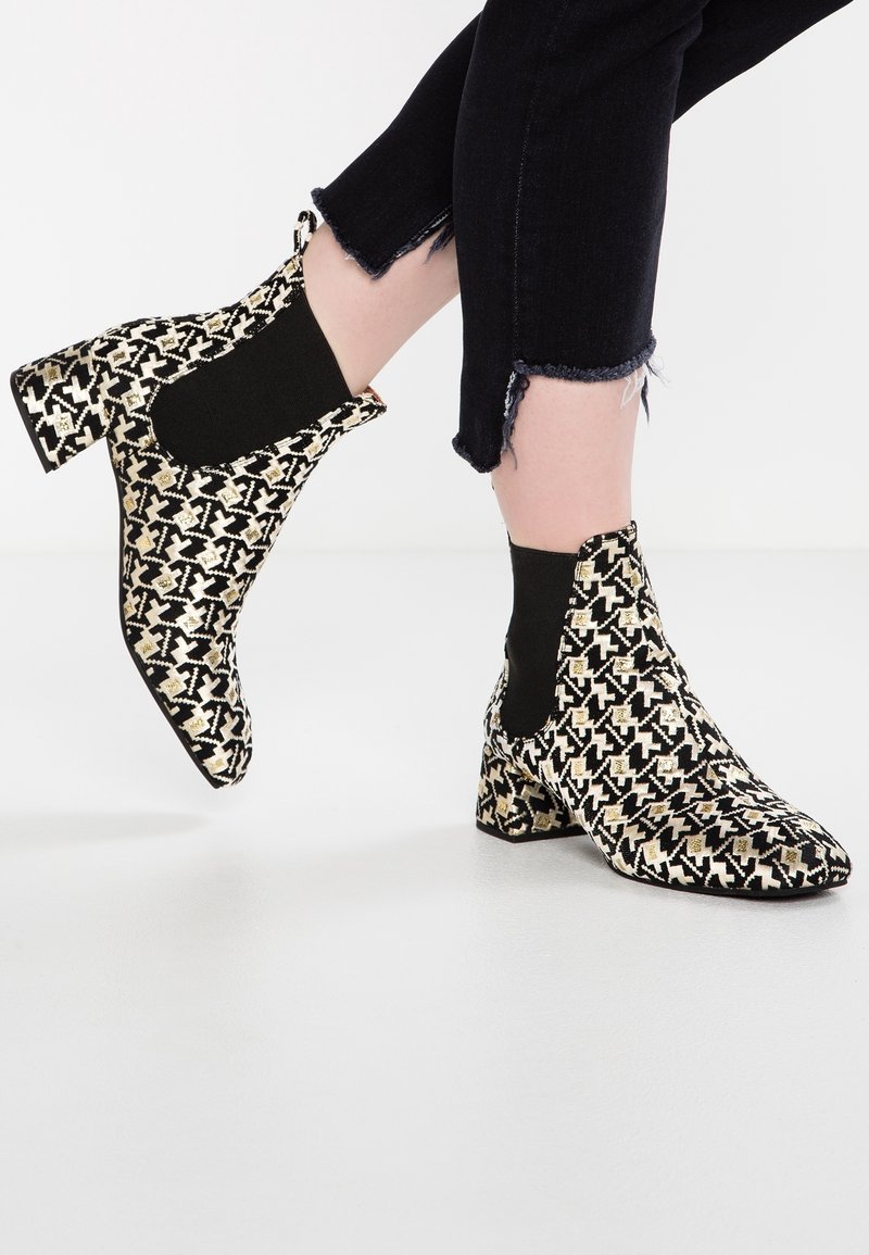 Bottines noires et dorées à motifs avec talons carrés portées avec un jean noir à ourlet effiloché sur fond blanc.