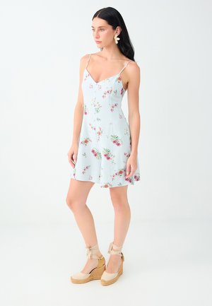 Femme aux longs cheveux foncés porte une robe fleurie bleu clair à fines bretelles et des sandales compensées beiges avec liens à la cheville sur fond blanc uni.