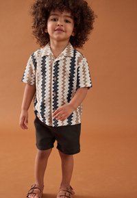 Kurzärmeliges Hemd mit einem Zickzack-Muster in Beige, Braun und Marineblau. Kombiniert mit dunklen Shorts. Steht vor einem braunen Hintergrund.