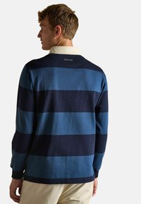 Mann mit dem Rücken zur Kamera, der ein blau- und navygestreiftes langärmliges Rugby-Shirt mit weißem Kragen und beigen Cordhose trägt.