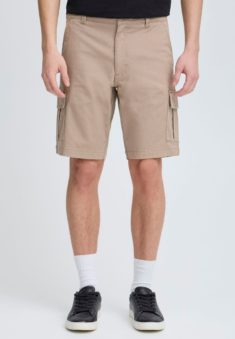 Pantaloni scurți cargo bej din bumbac, având buzunare laterale, o croială lejeră și un tiv drept. Purtați cu pantofi negri și șosete albe.