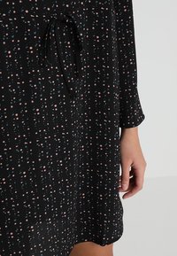 Musta mekko, jossa on pieniä pinkkejä ja sinisiä laikkuja, pitkät hihat ja ohut solmio vyötäröllä; oikea käsi näkyy rentona kyljessä.