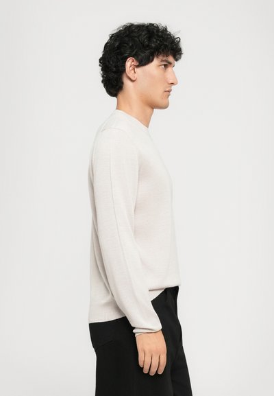 Mann mit lockigem, dunklem Haar, der einen hellbeigen Pullover mit langen Ärmeln und schwarze Hosen trägt, im Profil vor einem schlichten weißen Hintergrund stehend.