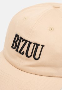 Beige baseballpet met zwarte geborduurde letters die "BIZUU" spellen op het voorpaneel.