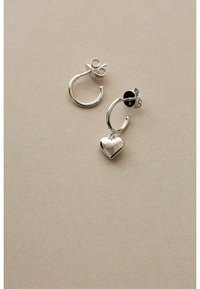 Orecchini a cerchio in argento con ciondolo a forma di cuore. Texture liscia, forma circolare e chiusura a perno. Design semplice su uno sfondo beige.