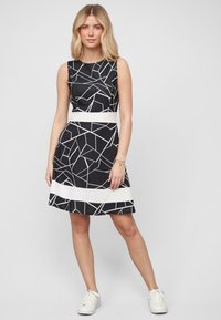 Robe noire avec un motif géométrique blanc, sans manches, taille cintrée et jupe évasée. Portée avec des baskets blanches.