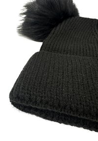 Berretto di maglia nera con texture a coste, caratterizzato da un grande pompon in pelliccia sintetica nera sulla sommità. Il materiale sembra morbido e caldo.