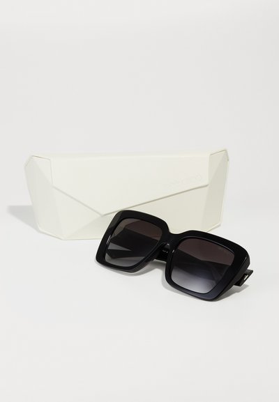 Schwarze übergroße Sonnenbrille mit Verlaufslinsen, ausgestattet mit einem dicken quadratischen Rahmen. Begleitet von einem geometrischen weißen Hartschalenetui.