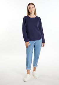 Dunkelblauer, gestrickter Pullover mit langen Ärmeln, kombiniert mit hellblauen, hochgekrempelten Jeans und weißen Sneakers. Einfache und lässige Gestaltung.