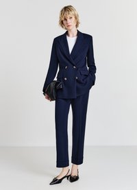 Blazer blu navy doppiopetto con due tasche frontali, pantaloni su misura abbinati e décolleté neri a punta. Tiene una borsa a mano nera.