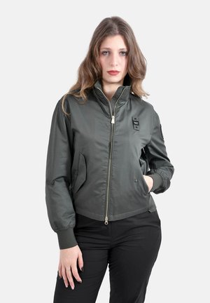 Donna con capelli lunghi che indossa una giacca bomber grigia con zip e pantaloni neri, in piedi con una mano in tasca su uno sfondo semplice.