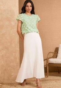 Top trasparente verde menta con applicazioni floreali testurizzate, abbinato a una gonna bianca fluida. Accentuato con sandali azzurri con cinturini.