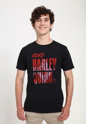 Ung mand smiler iført sort t-shirt med rød "XOXO HARLEY QUINN" tekst og lille hjerte på en ensartet lys baggrund.