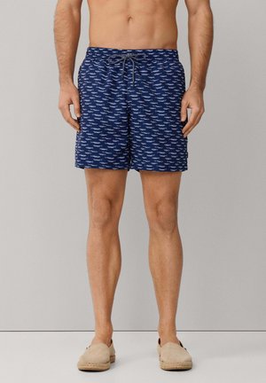 Torso et jambes d'homme portant un short de bain bleu marine à motif poissons blancs et des espadrilles beiges à enfiler sur un fond uni.