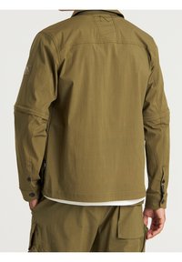 Mężczyzna ubrany w oliwkową koszulę z długim rękawem i pasujące spodnie cargo, widziany z tyłu, z rękami po bokach.