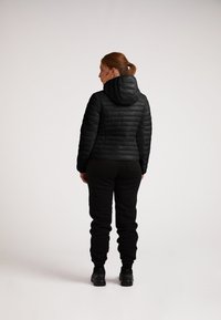 Giacca piumino nera con cappuccio, dalla texture trapuntata e design aderente, abbinata a pantaloni neri e calzature robuste.