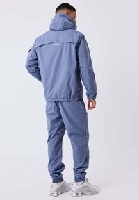 Man in een grijs-blauwe capuchonjack en bijpassende broek, met zijn rug naar de kijker gekeerd, met witte sneakers aan.