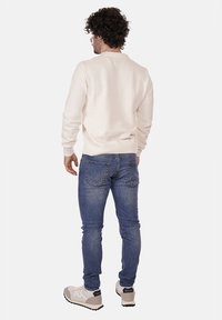 Pullover di maglia crema con collo a coste, abbinato a jeans di denim blu. Modella con scarpe da ginnastica beige; vista da dietro con i capelli ricci visibili.