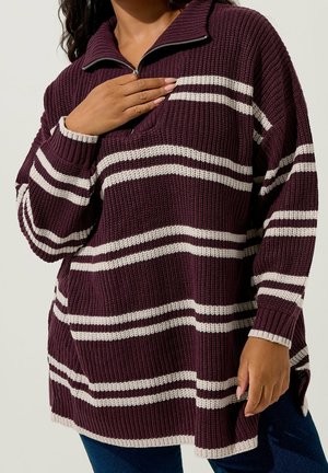 Femme portant un pull en maille épaisse avec des rayures blanches horizontales et une demi-fermeture éclair, visible du cou aux hanches.