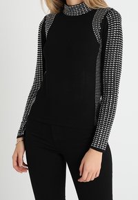 Musta pitkähihaine paita korkealla kauluksella, jossa on kontrastiväriin jäävillä kuviollisilla hihoilla, jotka on koristeltu pienillä valkoisilla pisteillä. Sileä pinta.