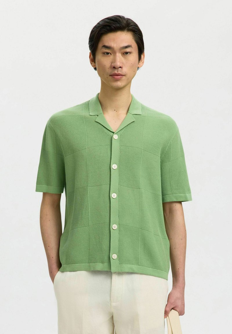 Homme portant une chemise tricotée à manches courtes vert clair avec boutons et pantalon crème, debout devant un fond clair uni.