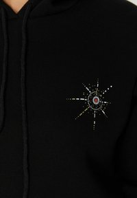 Sudadera negra con un diseño gráfico multicolor en el lado izquierdo del pecho, con patrones circulares y lineales en rojo, blanco, amarillo y plateado.