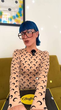 Person with blue hair, large square glasses, and crescent-patterned shirt sitting on green couch holding a black and yellow Buff box.