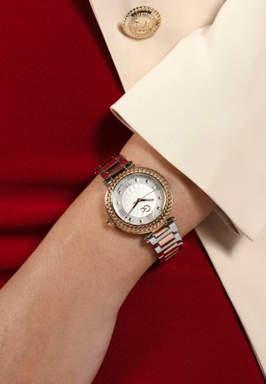 Reloj dorado con una esfera redonda de textura blanca, números romanos y una pulsera de metal bicolor, descansando sobre una manga beige clara contra un fondo rojo.