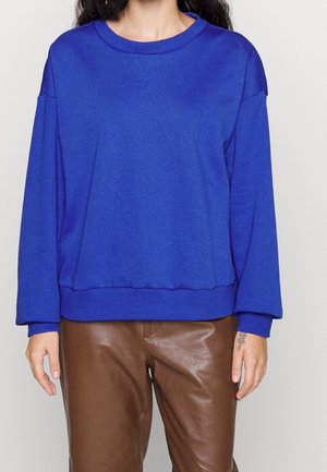 Personne portant un sweat-shirt à manches longues bleu roi associé à un pantalon en cuir marron, debout devant un fond clair uni.