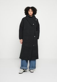 Black quilted coat featuring a high collar, asymmetrical closure, and button accents. Paired with wide-leg blue jeans and white shoes.